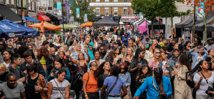 A lively street festival with a diverse crowd enjoying music and activities. Stalls and colorful umbrellas line the street under a clear sky.