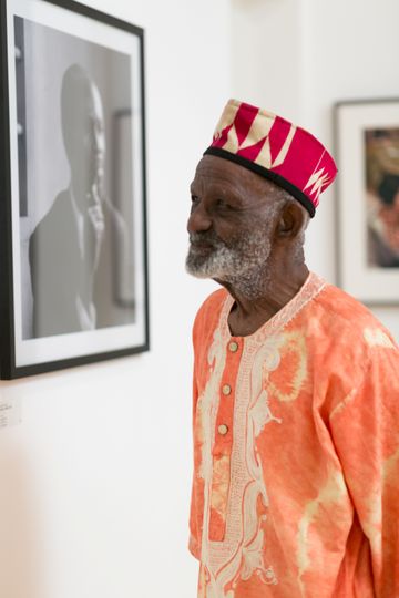 An elderly man in a vibrant orange African garment and hat admires black-and-white photography in an art gallery.