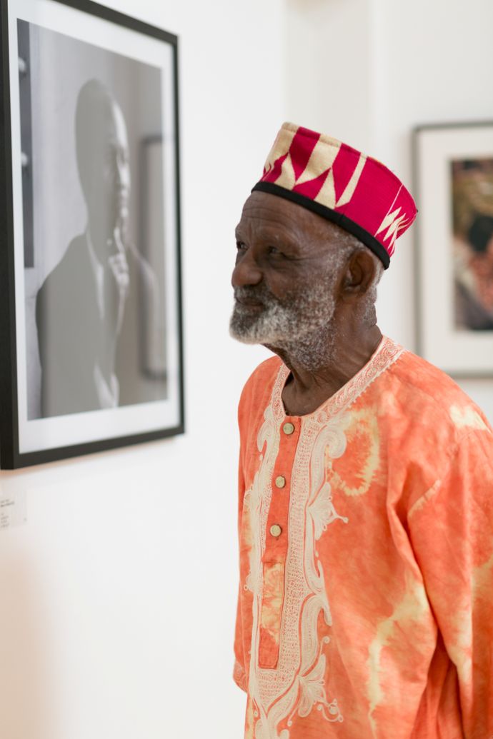 An elderly man in a vibrant orange African garment and hat admires black-and-white photography in an art gallery.