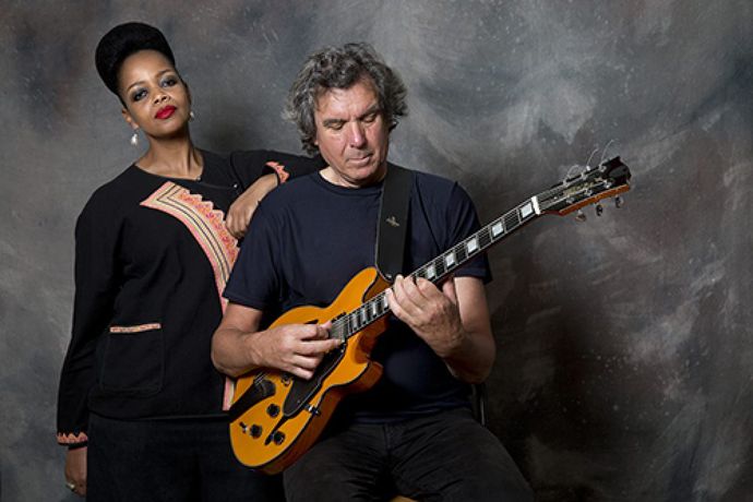 Two individuals standing besides each other playing an electric guitar, against a textured background.