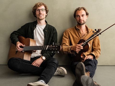 Two men sat down, one holding guitar the other holding violin