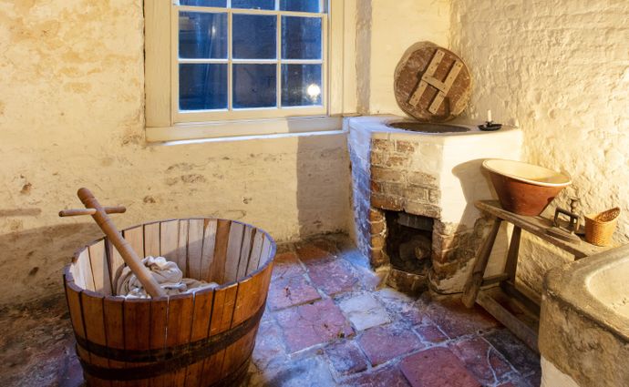Rustic washroom featuring wooden laundry tub, stone hearth, basin, candlelit walls, and window at dusk.