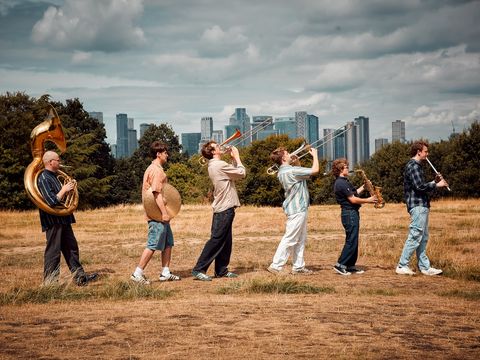 A brass band walking in a grassy field, playing instruments. City skyline and cloudy sky in the background.