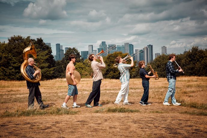 A brass band walking in a grassy field, playing instruments. City skyline and cloudy sky in the background.