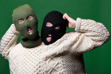Two people wearing knit balaclavas, one green and one black, make expressive faces against a green background, dressed in cream sweaters.