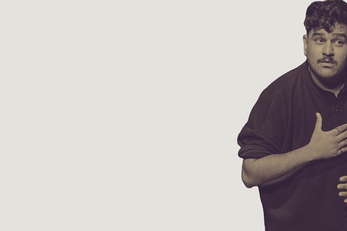 A person stands against a plain light‑coloured background with one hand placed on their chest and their body turned slightly to the side.