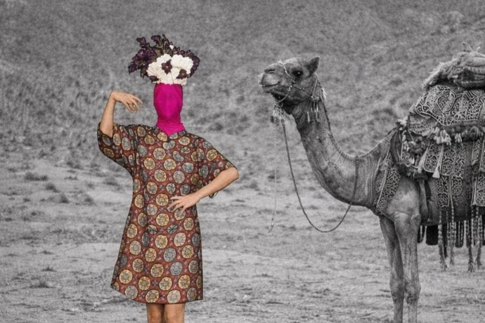 Person in a colorful patterned dress with a pink head covering and flowers, standing beside a decorated camel in a desert landscape.
