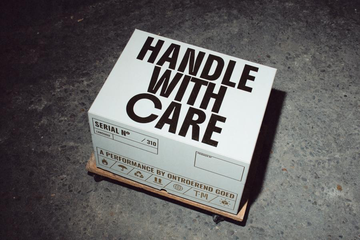 A white box labeled “HANDLE WITH CARE” rests on a wooden pallet on a concrete floor,