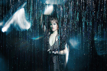 A performer stands behind a shimmering curtain under dramatic lighting, partially revealed in a stage-like setting.