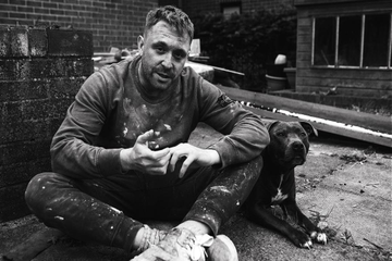 A person sits on the ground in an outdoor area covered in paint splatters, with a dog lying beside them.