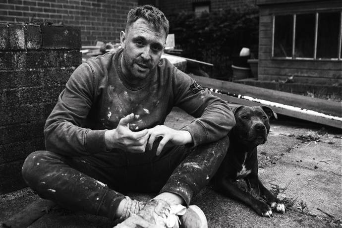 A person sits on the ground in an outdoor area covered in paint splatters, with a dog lying beside them.