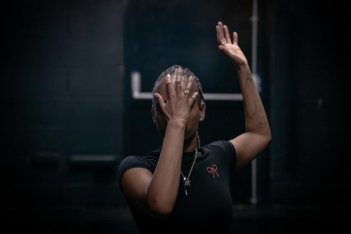 A person stands in a dimly lit space with one hand covering their face and the other arm raised above their head.