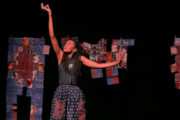A performer poses on a dark stage in a patterned outfit, framed by hanging patchwork fabric panels behind them.