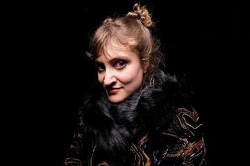 A softly lit portrait shows a person in a dark coat with a fur collar against a black background.