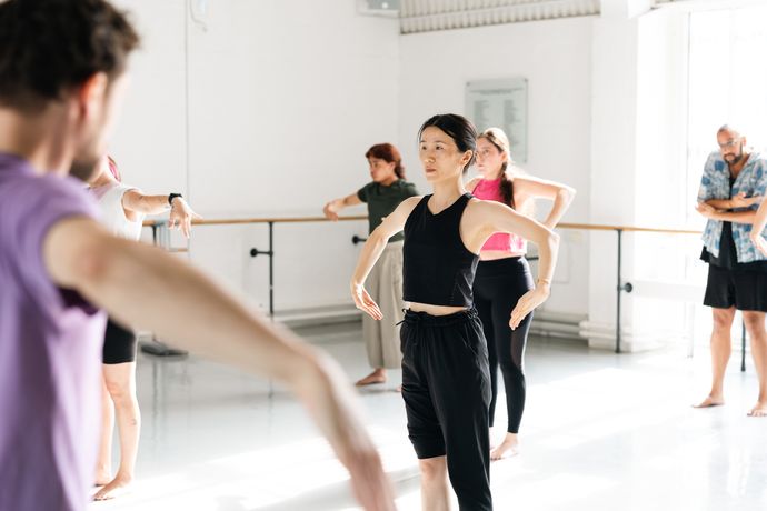 A group of people in casual fitness attire participate in a dance or movement class in a bright studio.