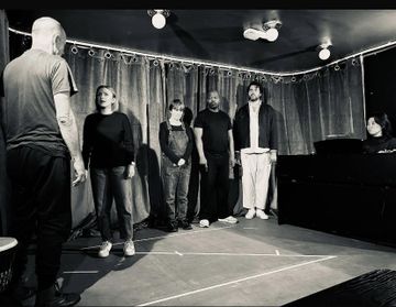 Black-and-white photo of several people standing in a line on a small stage or rehearsal space, facing another person at the left, with curtains behind them, overhead lights above, and tape markings visible on the floor.