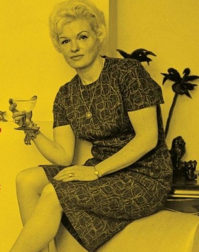 Woman with short blonde hair sits holding a cocktail glass, wearing a patterned dress, with tropical decor in the background, tinted yellow.