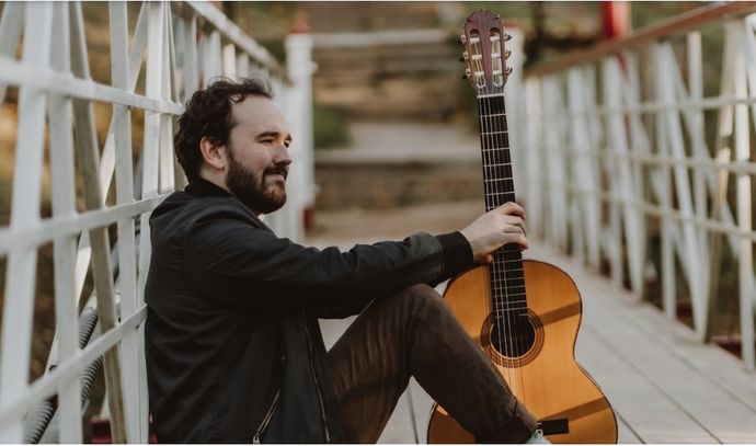 A person sits on a wooden bridge holding an acoustic guitar upright beside them.