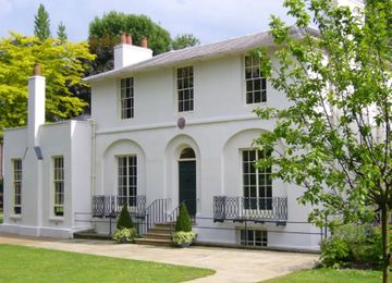 A two-story white house with arched windows, a central door, chimney stacks, and a small garden with trees and grass.