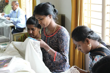 Women are focused on stitching intricate patterns on fabric in a well-lit room, with a man sewing in the background.
