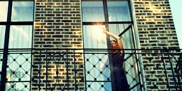 Person standing on a balcony, reaching up with a light source, against a brick wall with large windows.