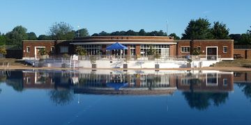 A brick building labeled "Cafe" with large windows and a blue tent is reflected in a calm pool under a clear blue sky.