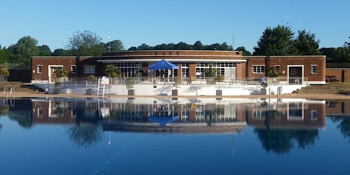 A brick building labeled "Cafe" with large windows and a blue tent is reflected in a calm pool under a clear blue sky.