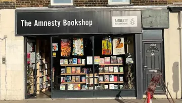 Front view of The Amnesty Bookshop with large glass windows displaying books and posters, and a black sign above reading 'The Amnesty Bookshop' alongside the Amnesty International logo