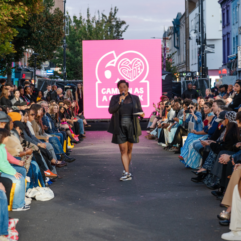 A fashion show on a city street with a large pink screen displaying "Camden 60 Years" and an audience seated on both sides of the runway.