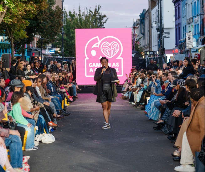 A fashion show on a city street with a large pink screen displaying "Camden 60 Years" and an audience seated on both sides of the runway.