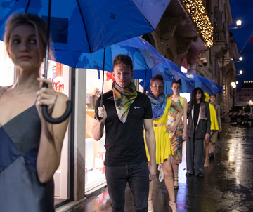 Models walk in a line on a wet street, holding blue umbrellas. They wear colorful outfits and scarves, with lights illuminating the scene.
