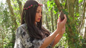 Woman in a floral dress and rose headband holds a pine cone, standing in a sunlit forest with ivy-covered trees.
