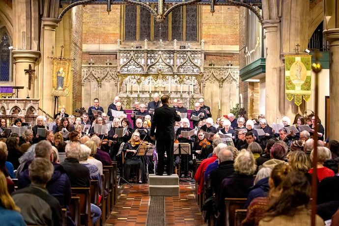 A choir and orchestra perform in a church with an audience seated. The conductor stands in front of the ensemble, with ornate architecture in the background.