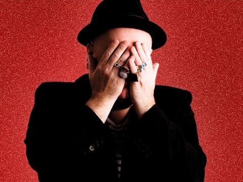 Person in a black hat and striped shirt, covering their face with hands wearing rings, against a textured red background.