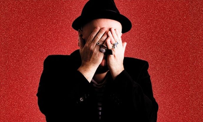 Person in a black hat and striped shirt, covering their face with hands wearing rings, against a textured red background.