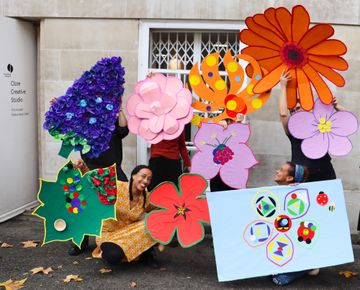 Four people holding large, colorful paper flowers in front of a building, smiling and crouching on a street covered with fallen leaves.
