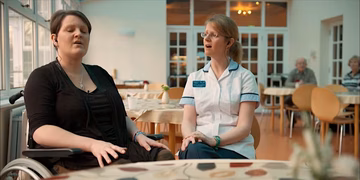 woman in wheelchair wearing earphones closes her eyes, sitting next to woman in white nurses uniform. they both have their mouths open in singing pose