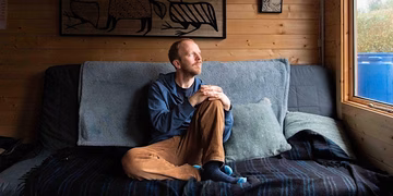 Man sits on sofa bed with blankets and cushions on with wooden panelling behind, looking out of window with sunlight on face