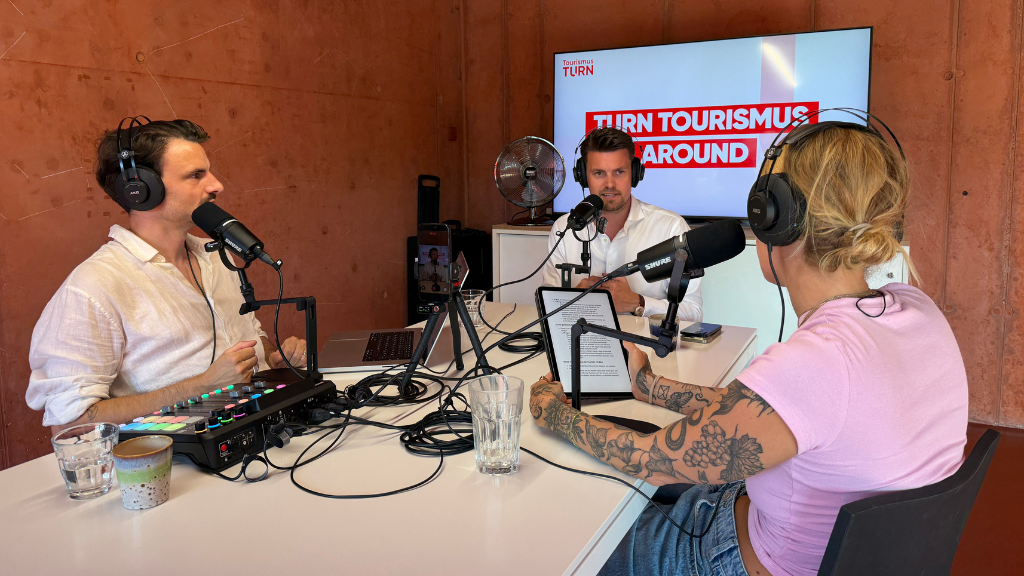 Drei Personen mit Mikrofonen und Kopfhörern sitzen an einem Tisch in einem Podcast-Studio, im Hintergrund ein Bildschirm mit der Aufschrift 'TURN TOURISMUS AROUND'