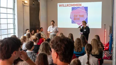 Maren und Marvin stehen vor einer Gruppe sitzender Menschen und sprechen vor einer Leinwand mit dem Schriftzug 'OUT OF THE BOX 2026'