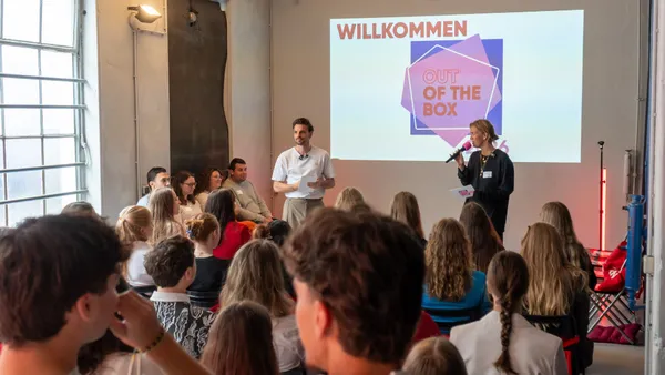 Maren und Marvin stehen vor einer Gruppe sitzender Menschen und sprechen vor einer Leinwand mit dem Schriftzug 'OUT OF THE BOX 2026'