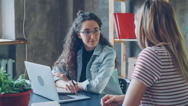 Zwei Frauen sitzen an einem Tisch, eine mit lockigem Haar und Jeansjacke vor einem Laptop, die andere mit gestreiftem T-Shirt ihnen gegenüber