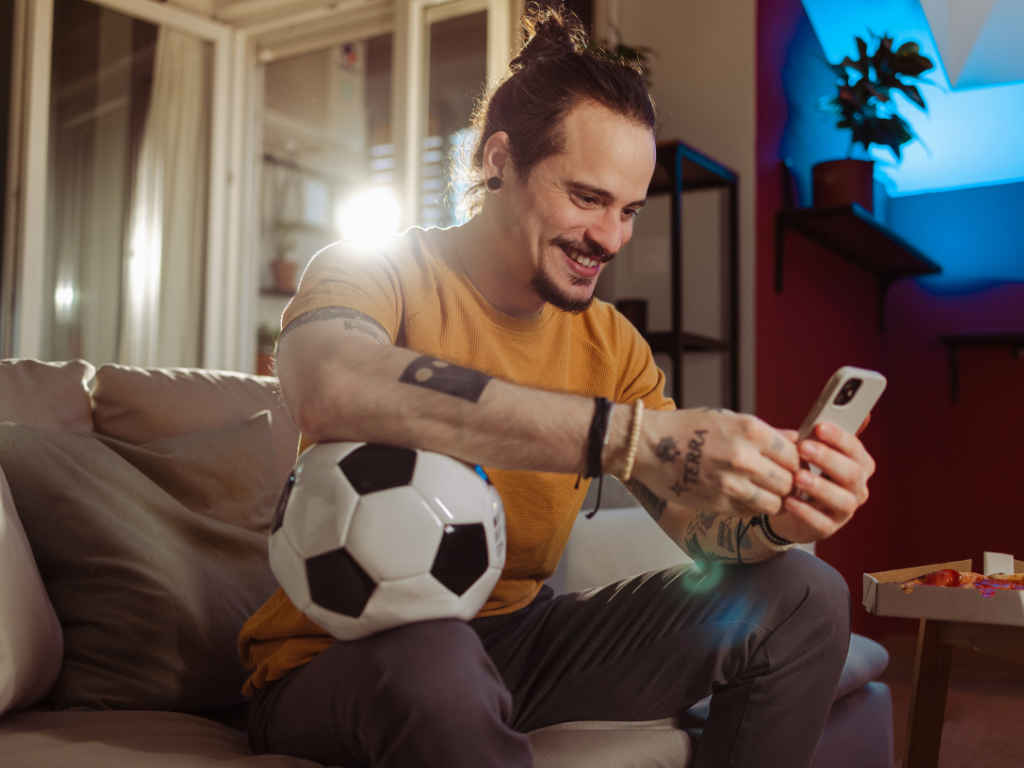 Ein Mann mit Tätowierungen und einem Haarknoten sitzt auf einer Couch, hält einen Fußball in der Hand und lächelt in einem warm beleuchteten Raum in sein Handy.