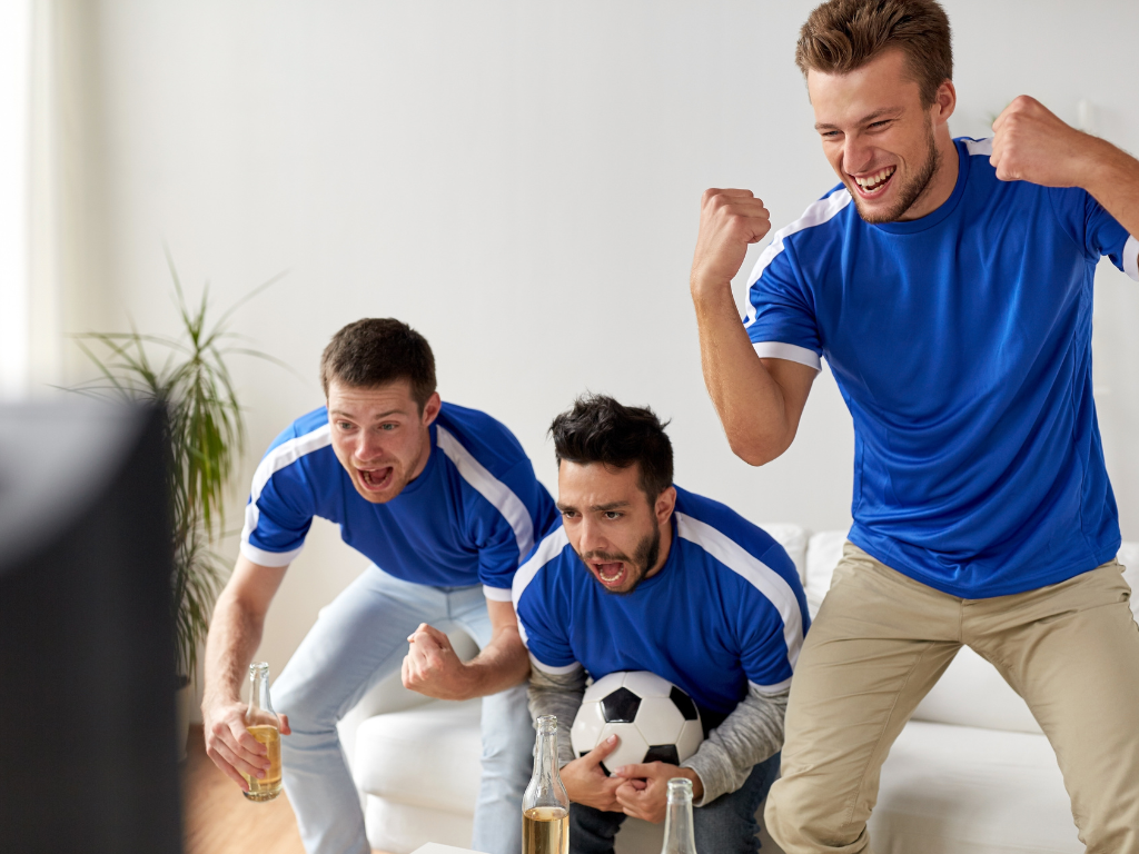 Drei begeisterte Männer in blauen Trikots jubeln, während sie ein Fußballspiel im Fernsehen verfolgen. Einer hält einen Fußball in der Hand, die anderen haben Glasflaschen in der Hand.