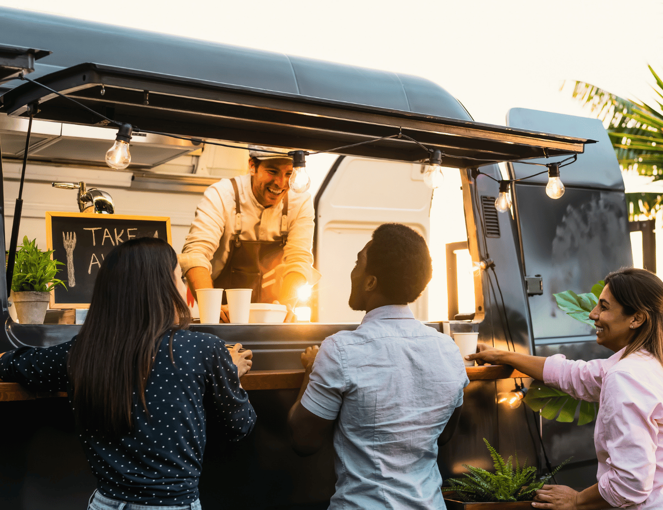 Ein Food-Truck-Verkäufer serviert drei lächelnden Kunden an einem Standort im Freien Getränke, mit einer kleinen Kreidetafel und hängenden Lichtern.