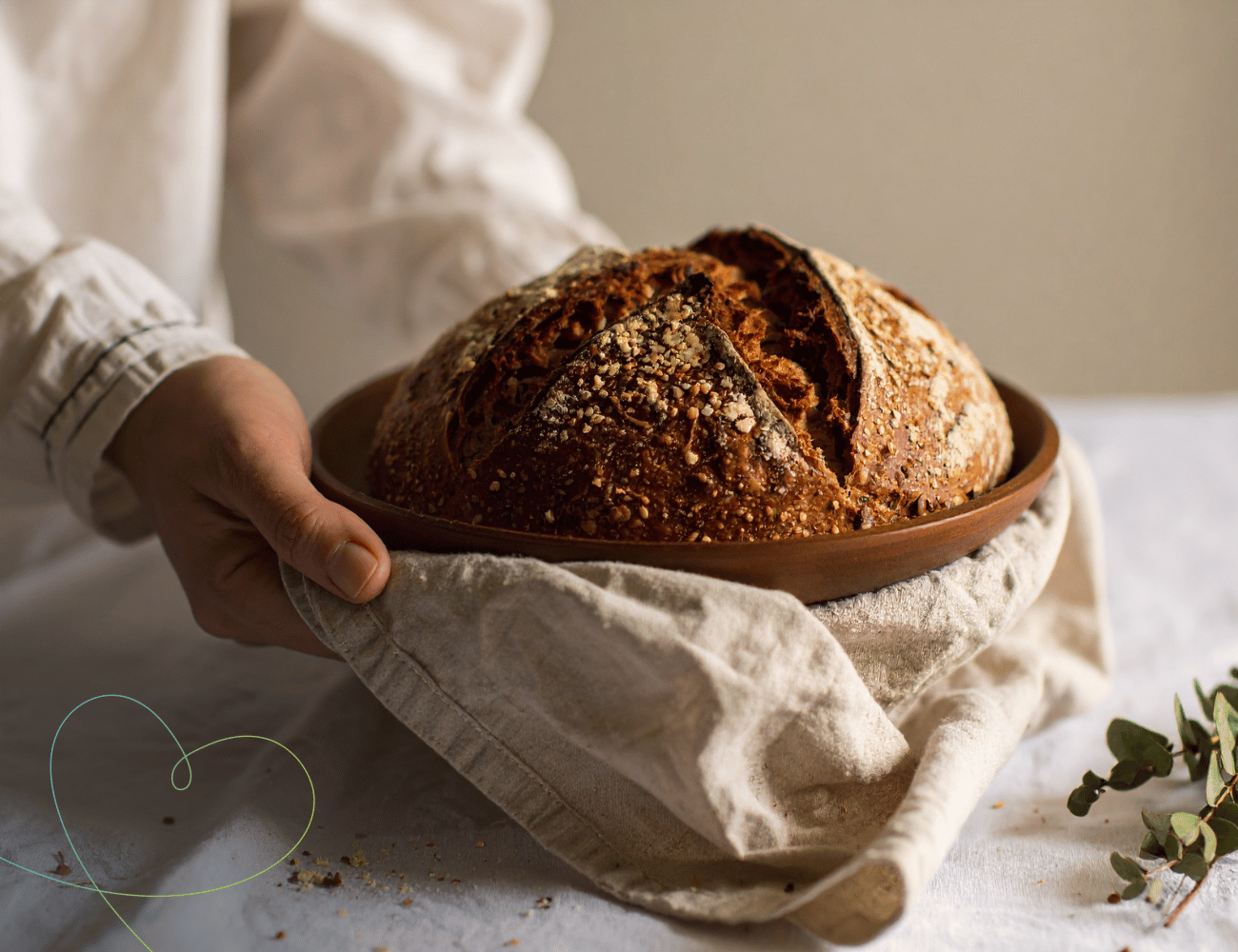 Hände, die einen runden, knusprigen Brotlaib in einer Holzschale halten, die in ein Leinentuch gewickelt auf einem hellen Tisch liegt.