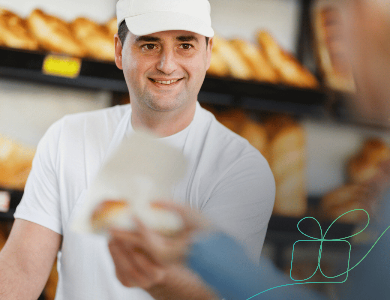 Ein lächelnder Bäcker mit weißer Mütze reicht einem Kunden in einer Bäckerei, in der Brote in Regalen ausliegen, ein verpacktes Gebäckstück.