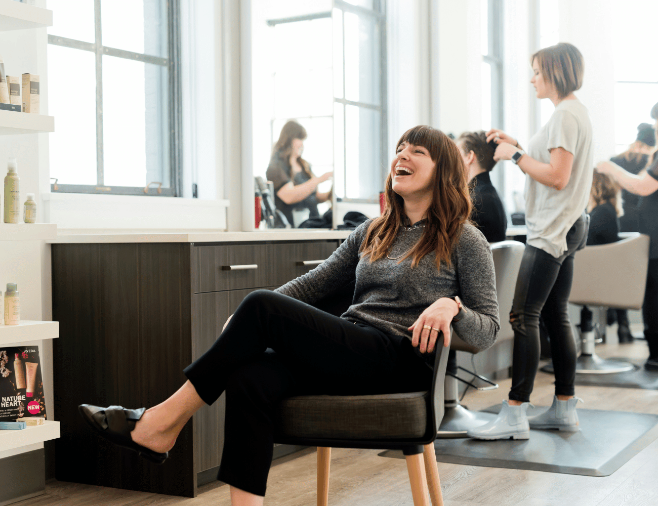 Eine Frau sitzt lachend in einem Friseurstuhl, während Stylist:innen im Hintergrund die Haare ihrer Kund:innen frisieren.