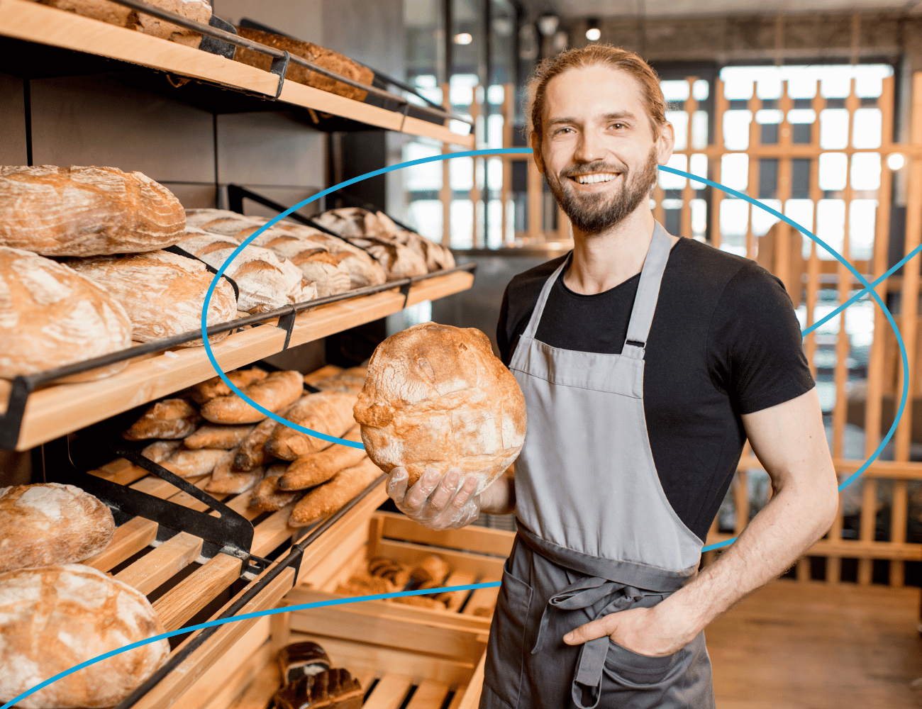 Lächelnder Bäcker in grauer Schürze, der einen runden Laib Brot in einem Bäckereigang hält, der mit Holzregalen mit frischem Brot gesäumt ist.