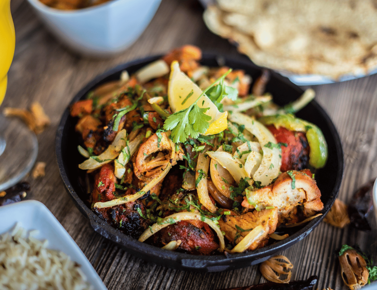 Ein knuspriges Gericht aus gegrilltem Hähnchenfleisch mit Zwiebeln und Kräutern, garniert mit einer Zitronenscheibe und Koriander, serviert in einer schwarzen Pfanne.